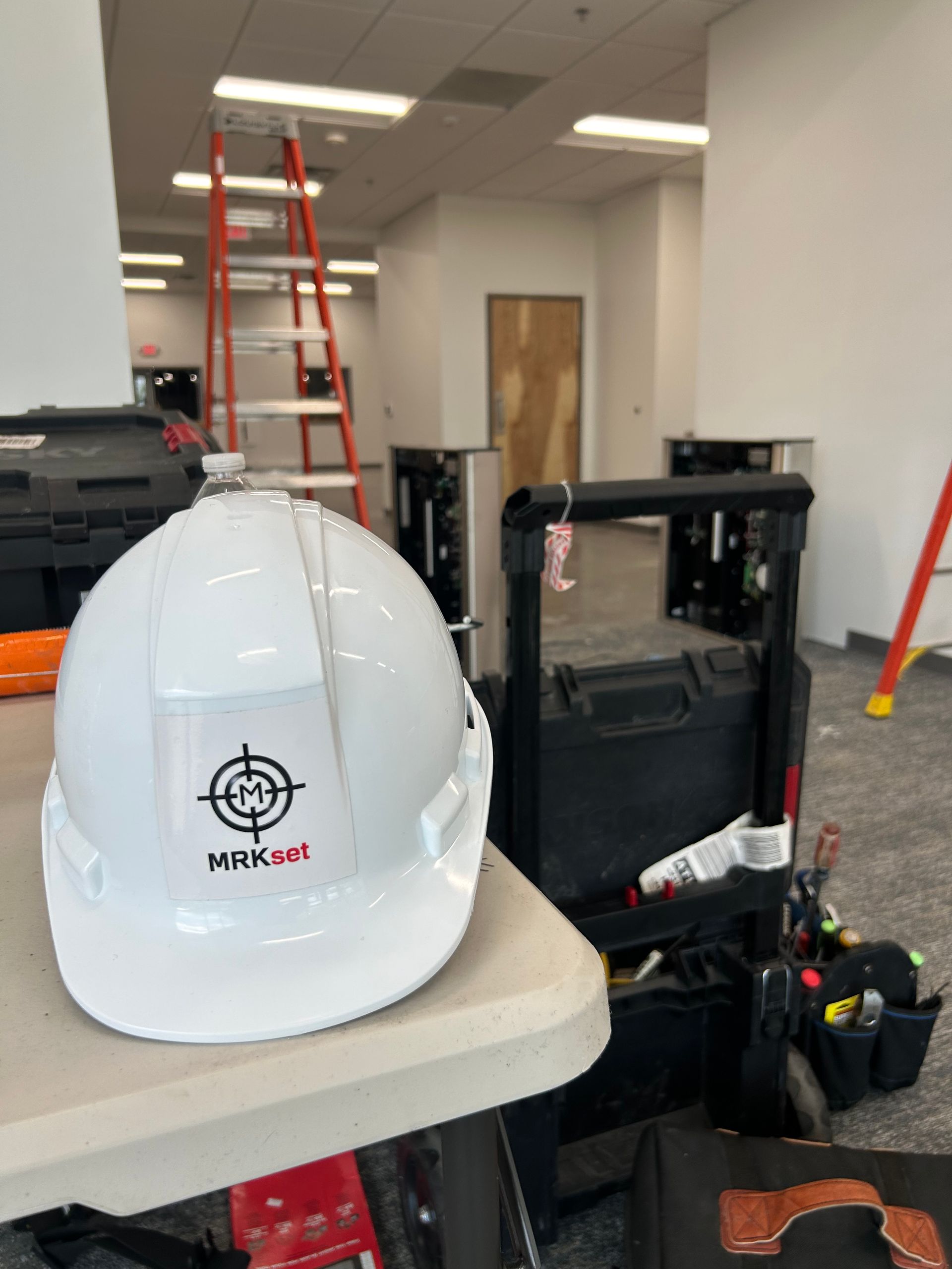 White hard hat with MRKset logo on table, tool bag and ladder in a hallway.