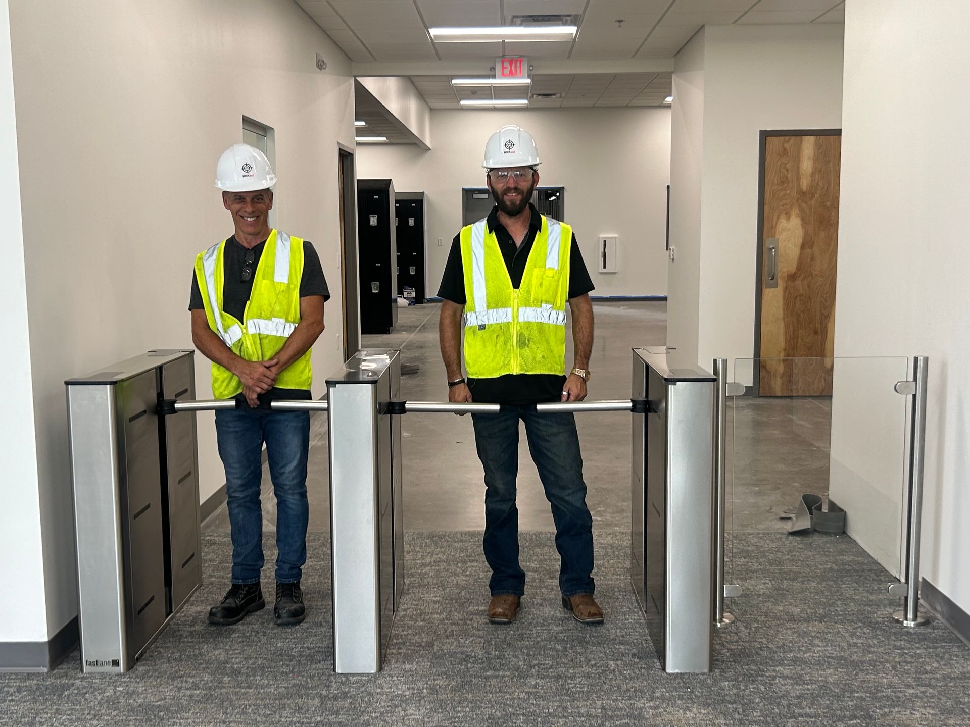 Two people in hard hats and vests stand in front of turnstiles in a hallway.