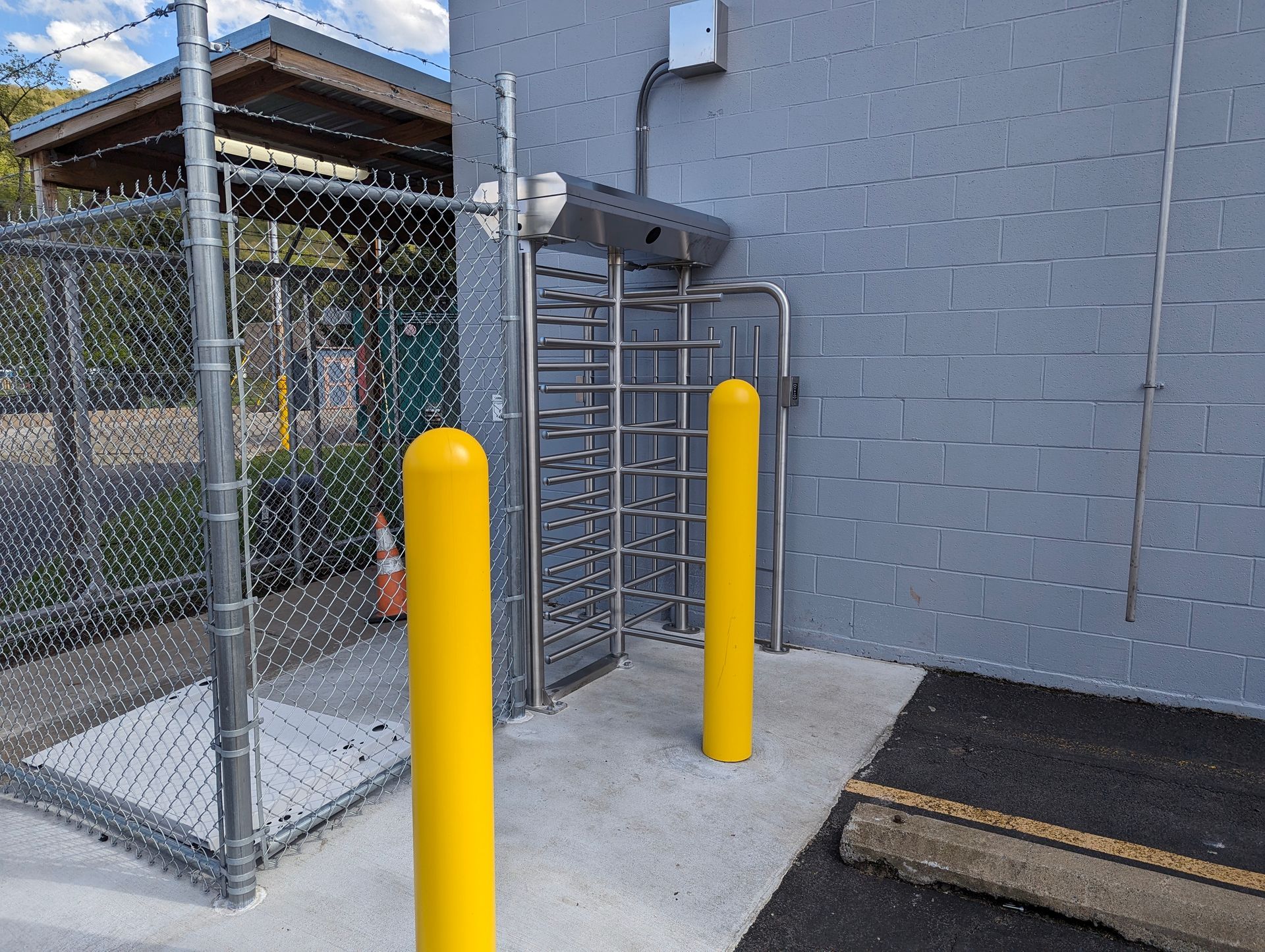 Turnstile entrance with yellow bollards, chain-link fence, and grey building.