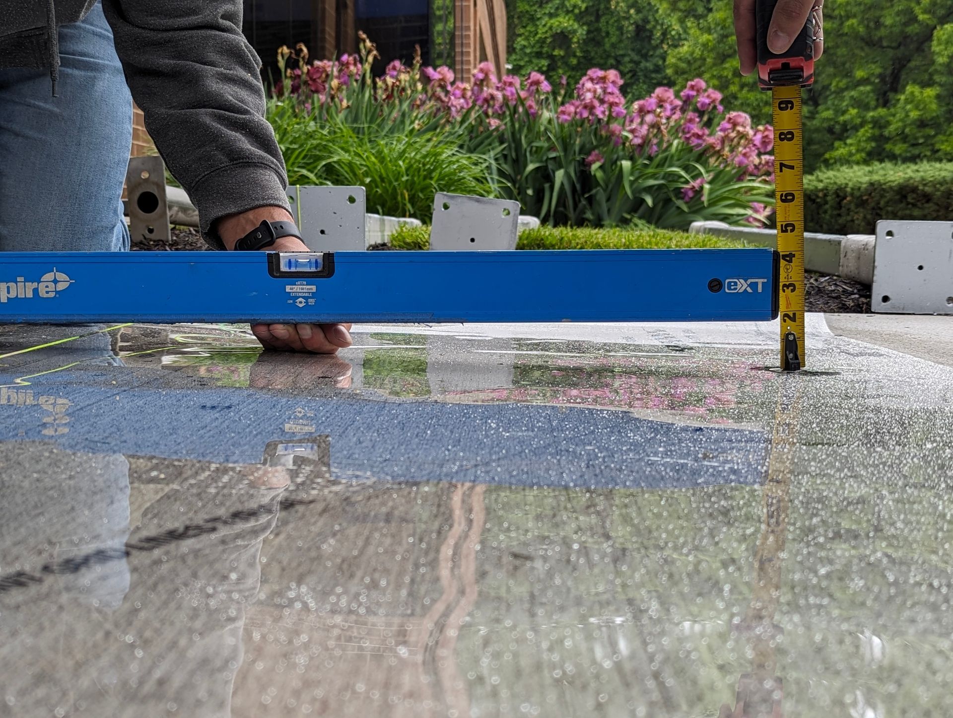 Person using a level and measuring tape on a wet, gray surface outdoors.
