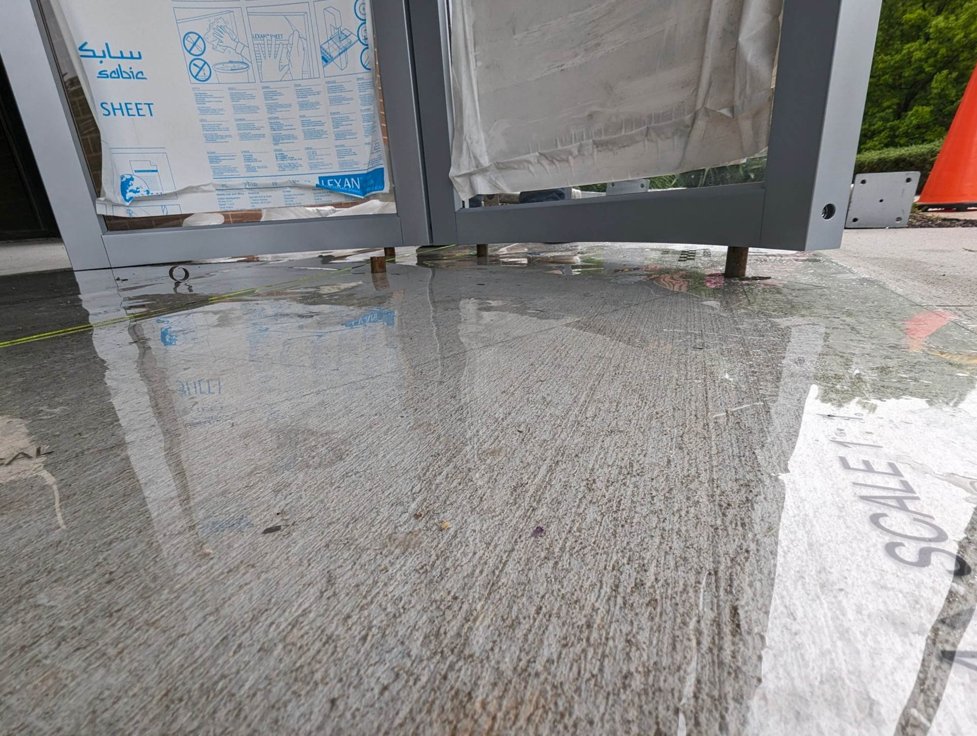 Water puddles on a concrete surface under a silver-framed structure.