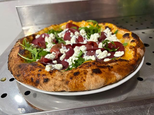 Pizza cotta nel forno a legna condita con verdure, fette di barbabietola e formaggio sbriciolato, servita su un vassoio di metallo.