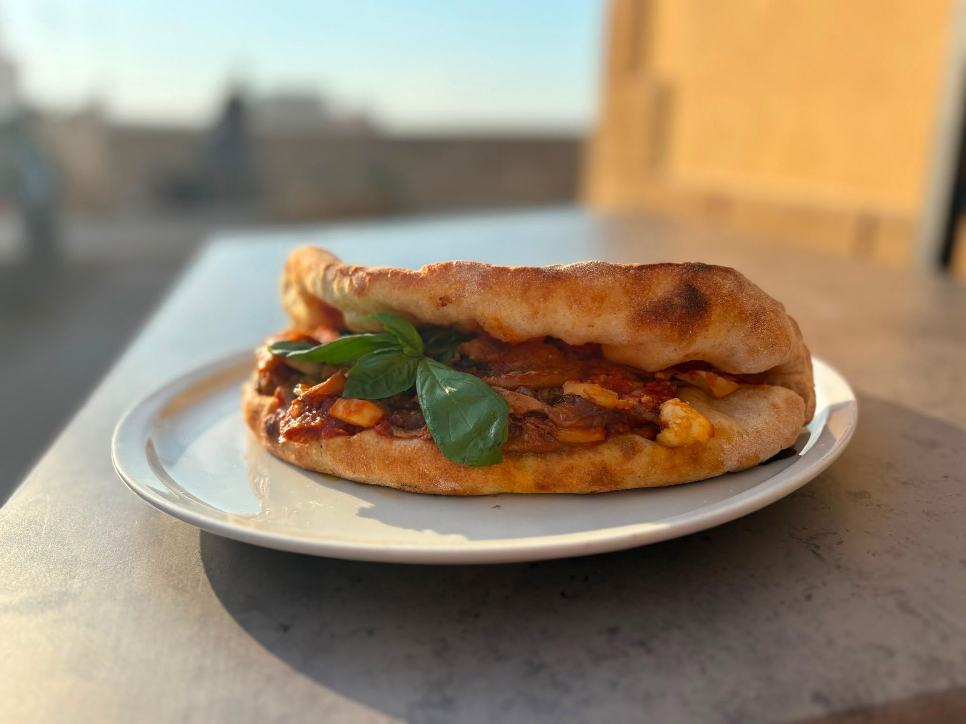 Un panino su un piatto bianco con foglie di basilico, appoggiato su un tavolo all'aperto sotto il caldo sole.