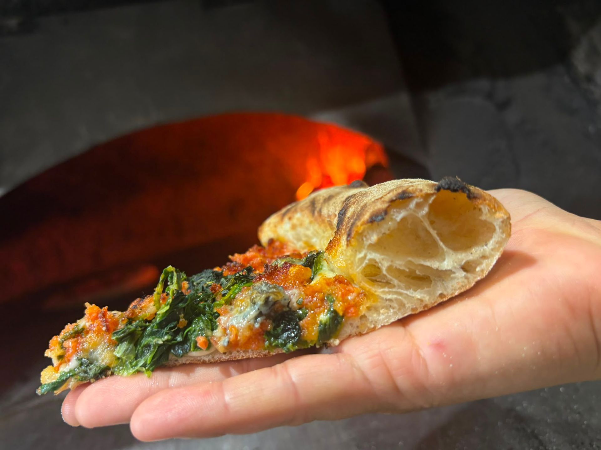 Una mano regge una fetta di pizza con formaggio fuso e verdure davanti a un forno a legna.