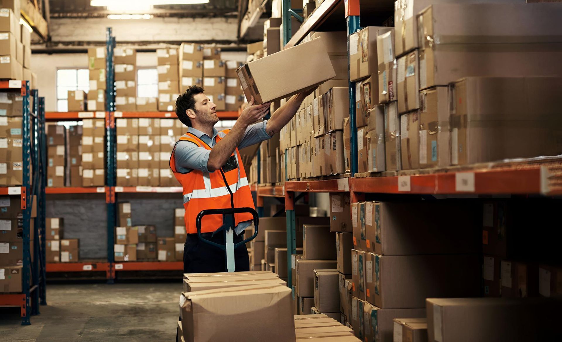 Warehouse worker in an orange vest reaching for a box on a shelf filled with packages.