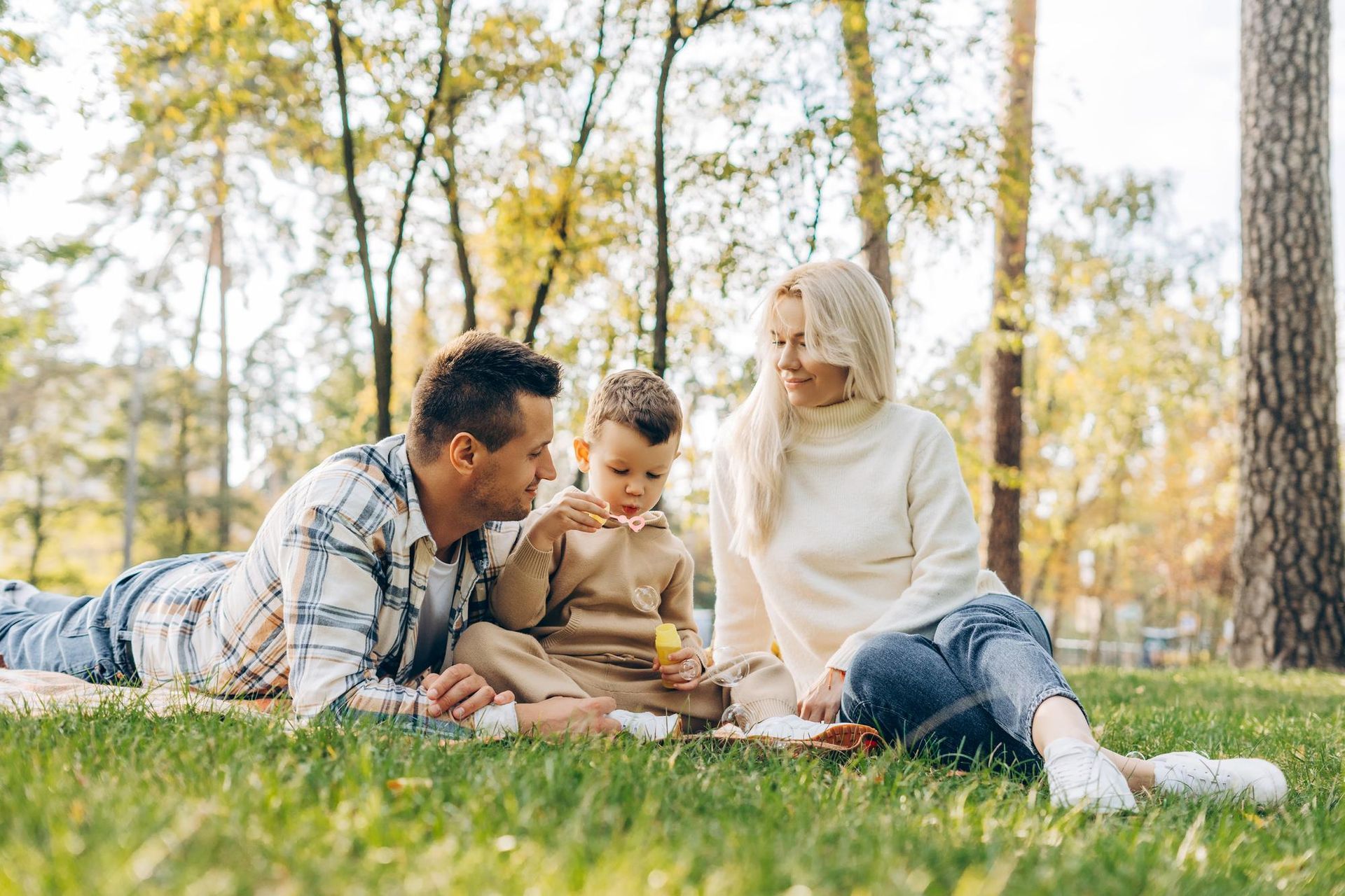 Family enjoying a picnic on a grassy lawn. Man and woman, child. Sunlight streams through trees.