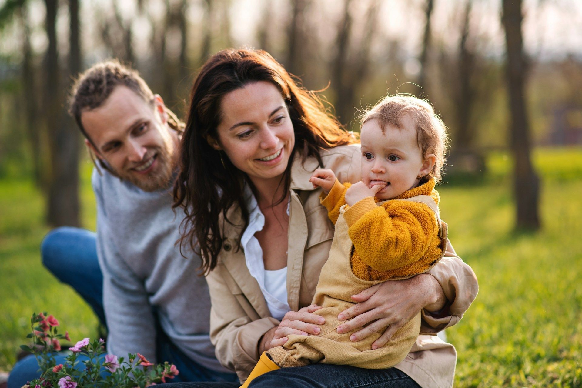 Family in a grassy park: parents smiling, holding a baby in yellow overalls. Trees in background.