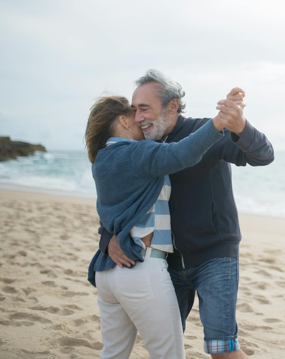 Couple dancing on a sandy beach; ocean in background. Man smiles, holds woman in embrace.
