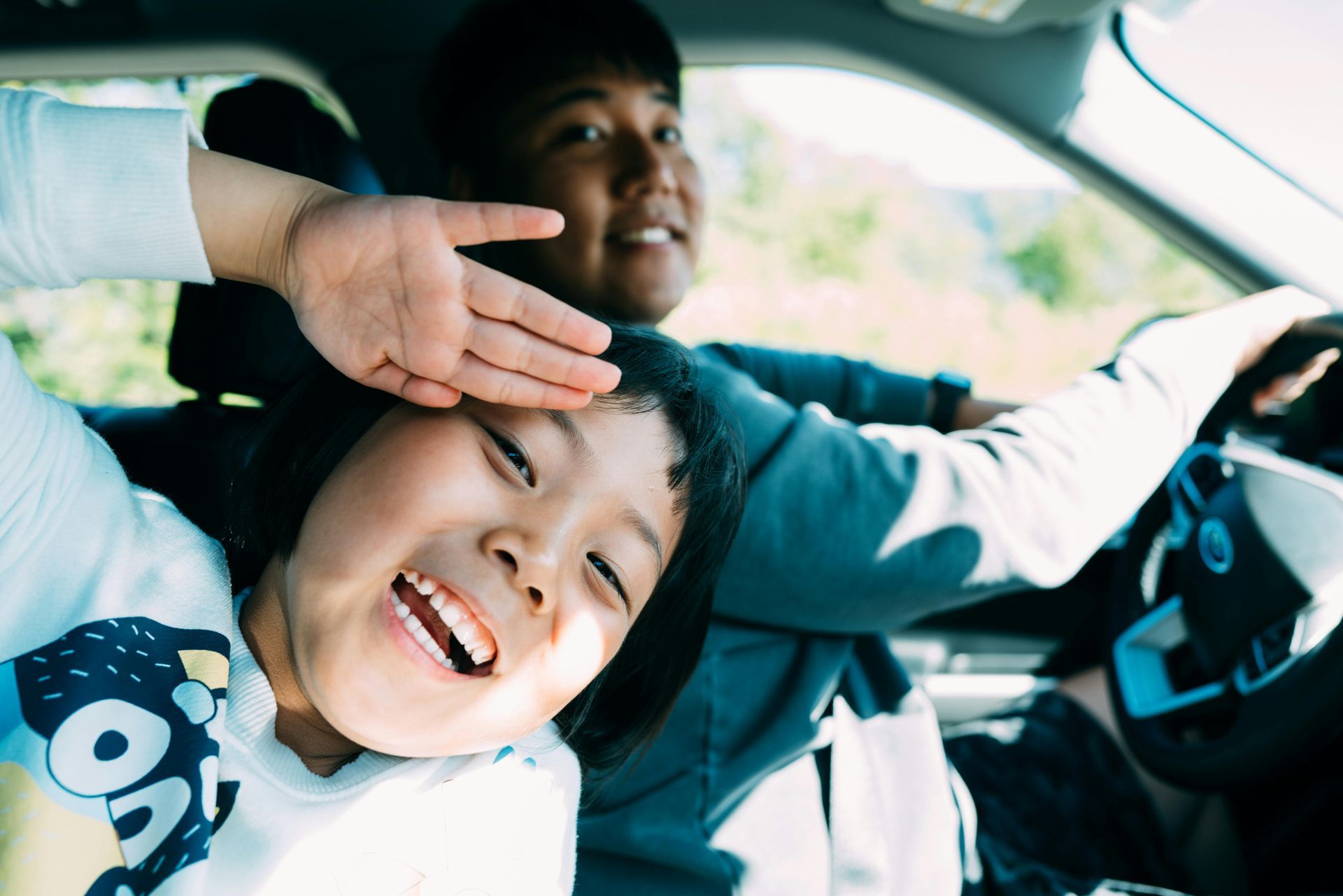 Child laughs, leaning towards the camera from car. Adult smiles while driving. Sunlight.