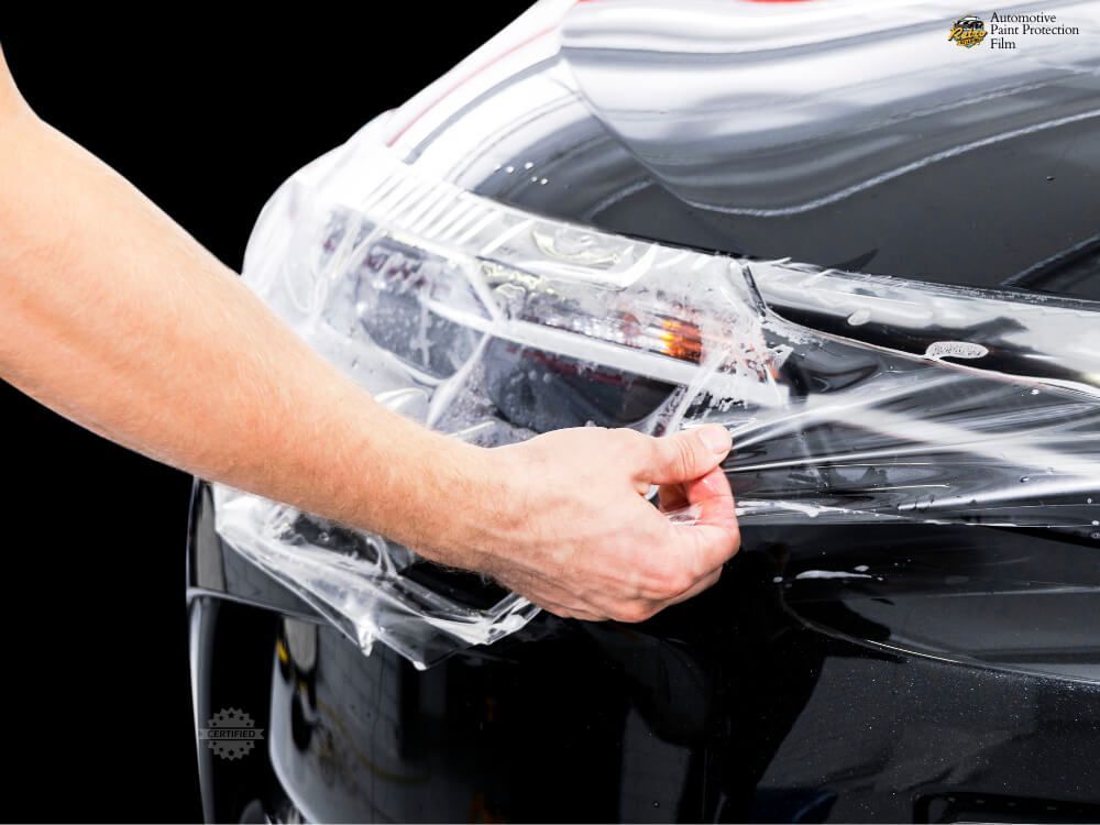 Hands applying clear protective film to the front of a black car hood