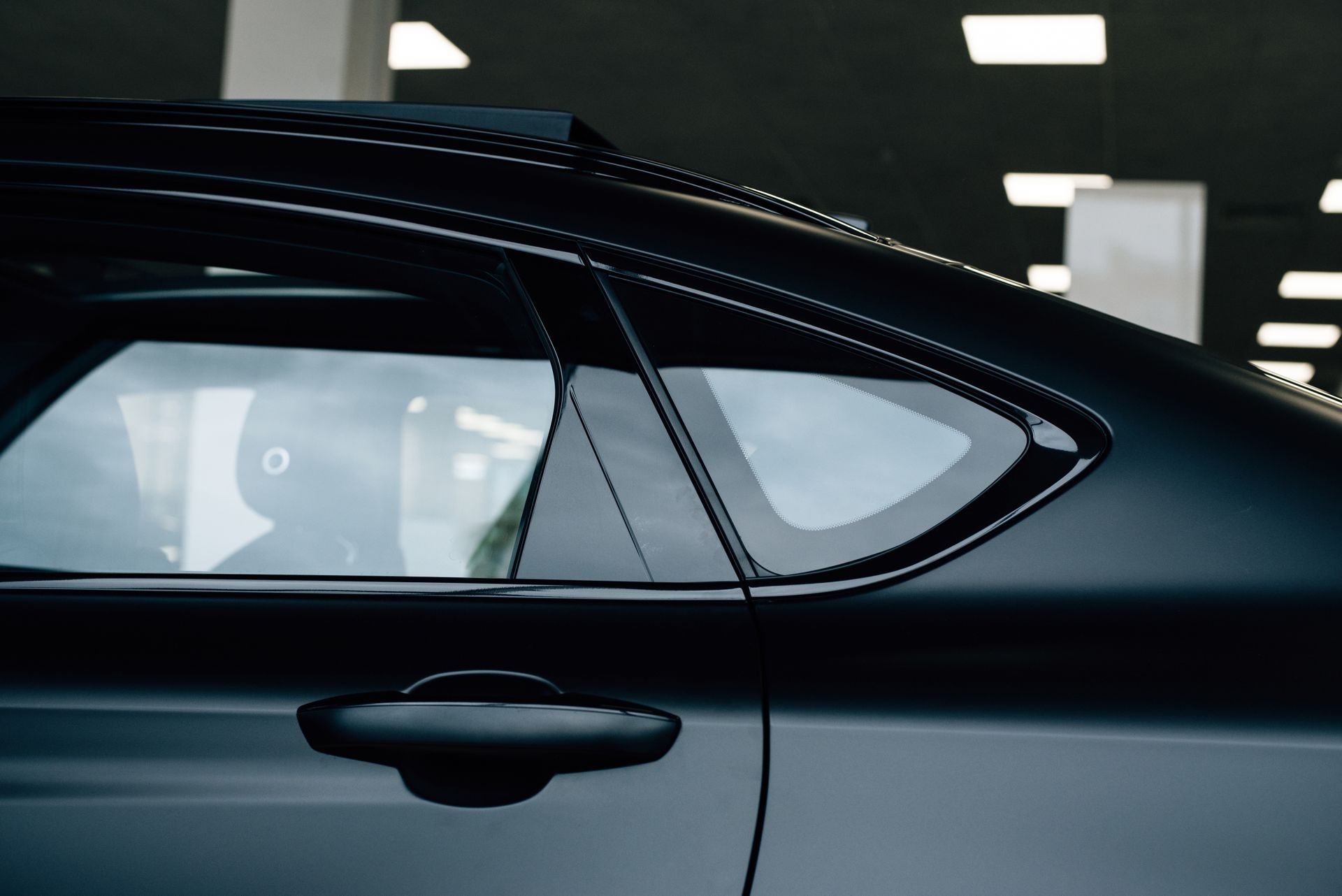 Side view of a black car door and rear window in a garage, with overhead lights reflected in the glass.