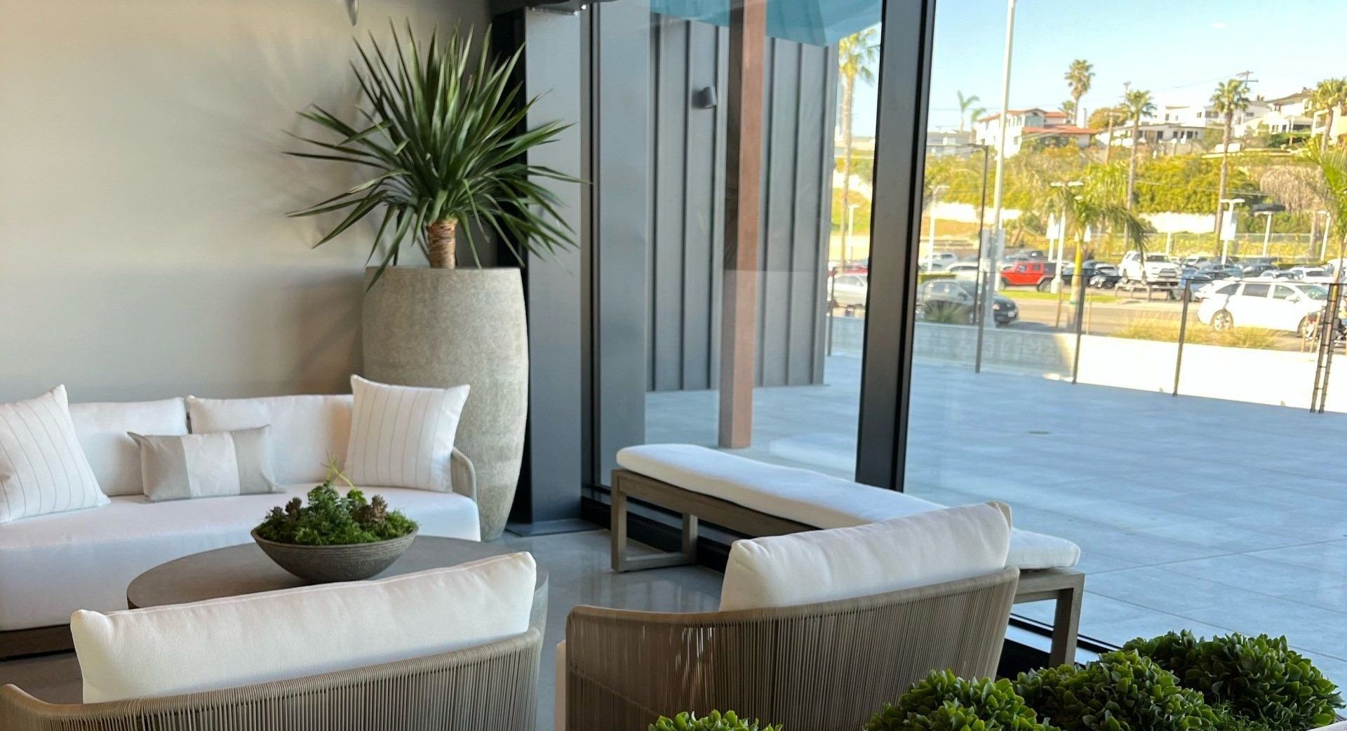 Modern outdoor patio lounge with white seating, potted plants, and glass walls overlooking a sunny view