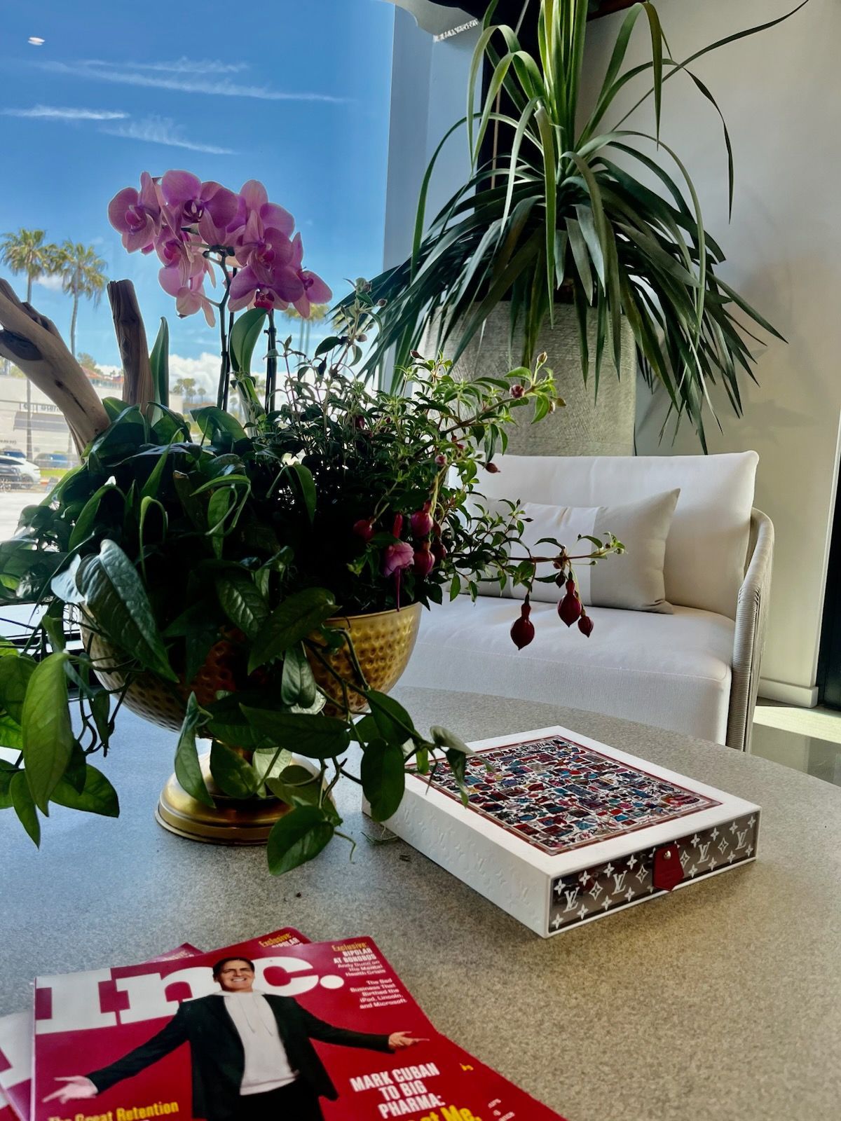 Living room table with orchids, a patterned box, magazines, and a white chair by a sunlit window