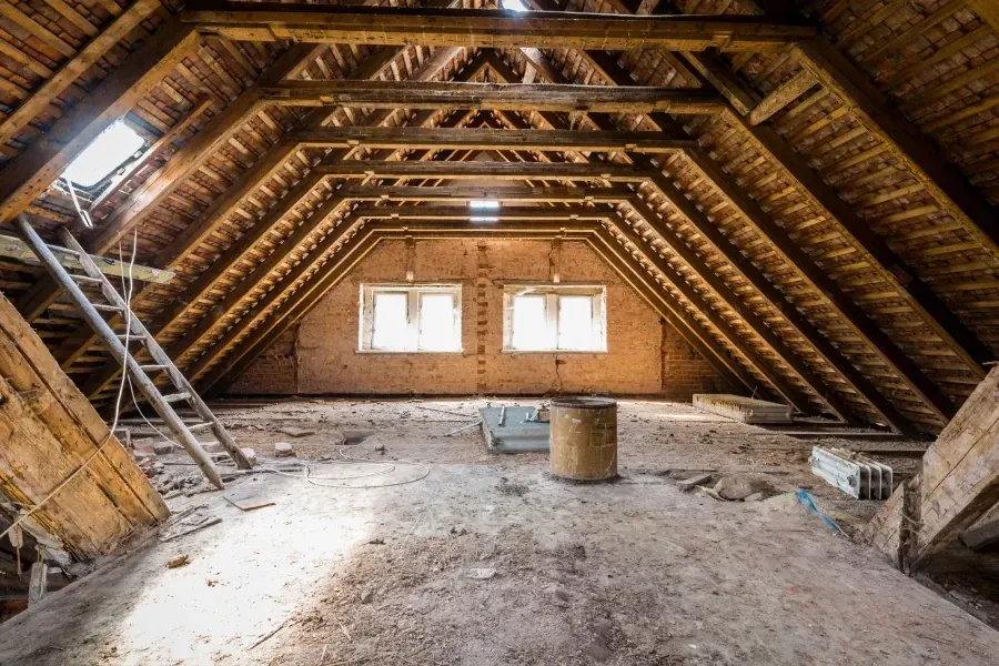An unfinished attic with exposed wooden rafters, a ladder on the left, two windows on the back wall, and dusty floor.