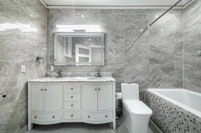 A modern bathroom featuring grey marble-tiled walls, a white double vanity, a framed mirror, and a white toilet.