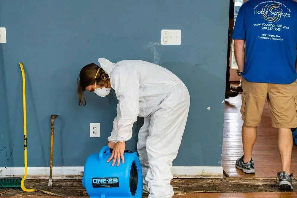 A person in a protective white suit and mask positions a blue air mover on a floor while another person stands nearby.