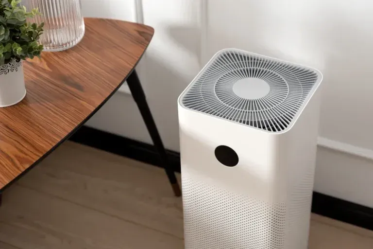 A white air purifier stands on a light-colored floor next to a small wooden table with a potted plant.