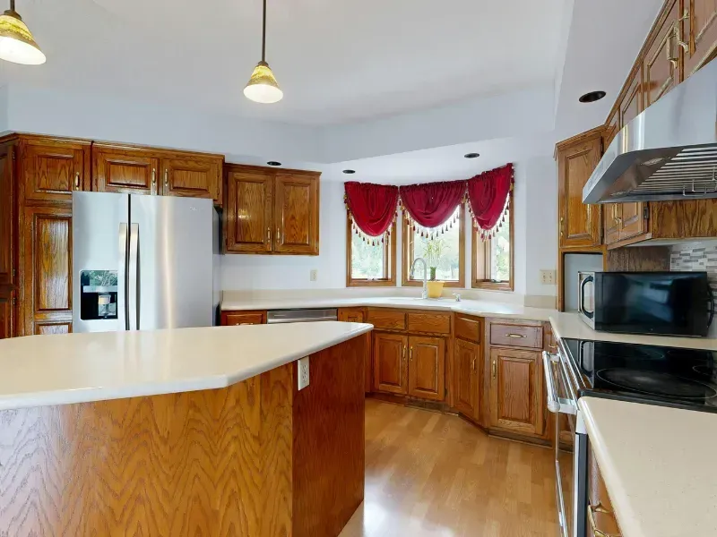 A bright kitchen with honey oak cabinets, white countertops, a stainless steel refrigerator, and windows with red valances.