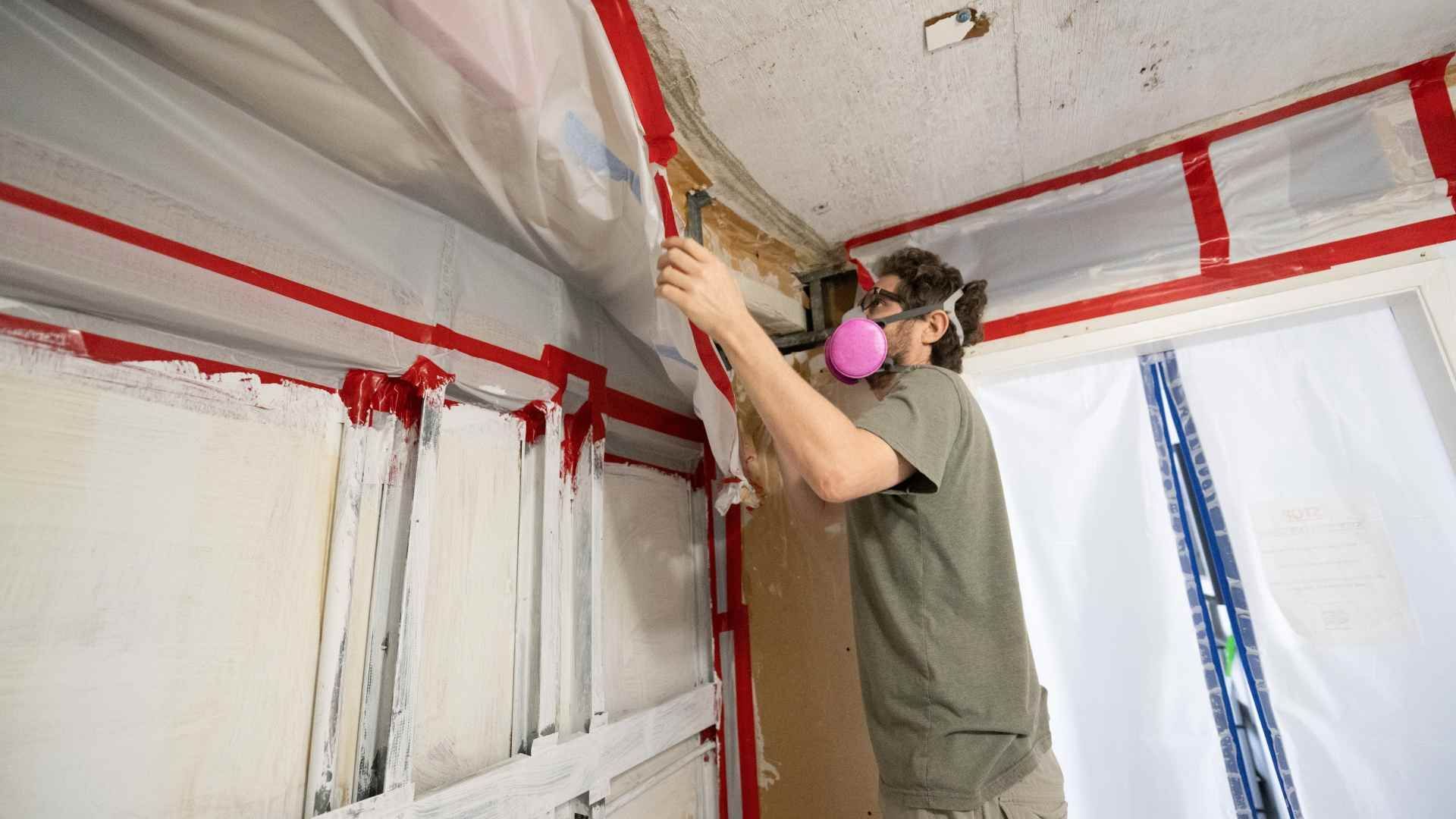 A person wearing a pink respirator mask tapes plastic sheeting over a wall during a home renovation project.