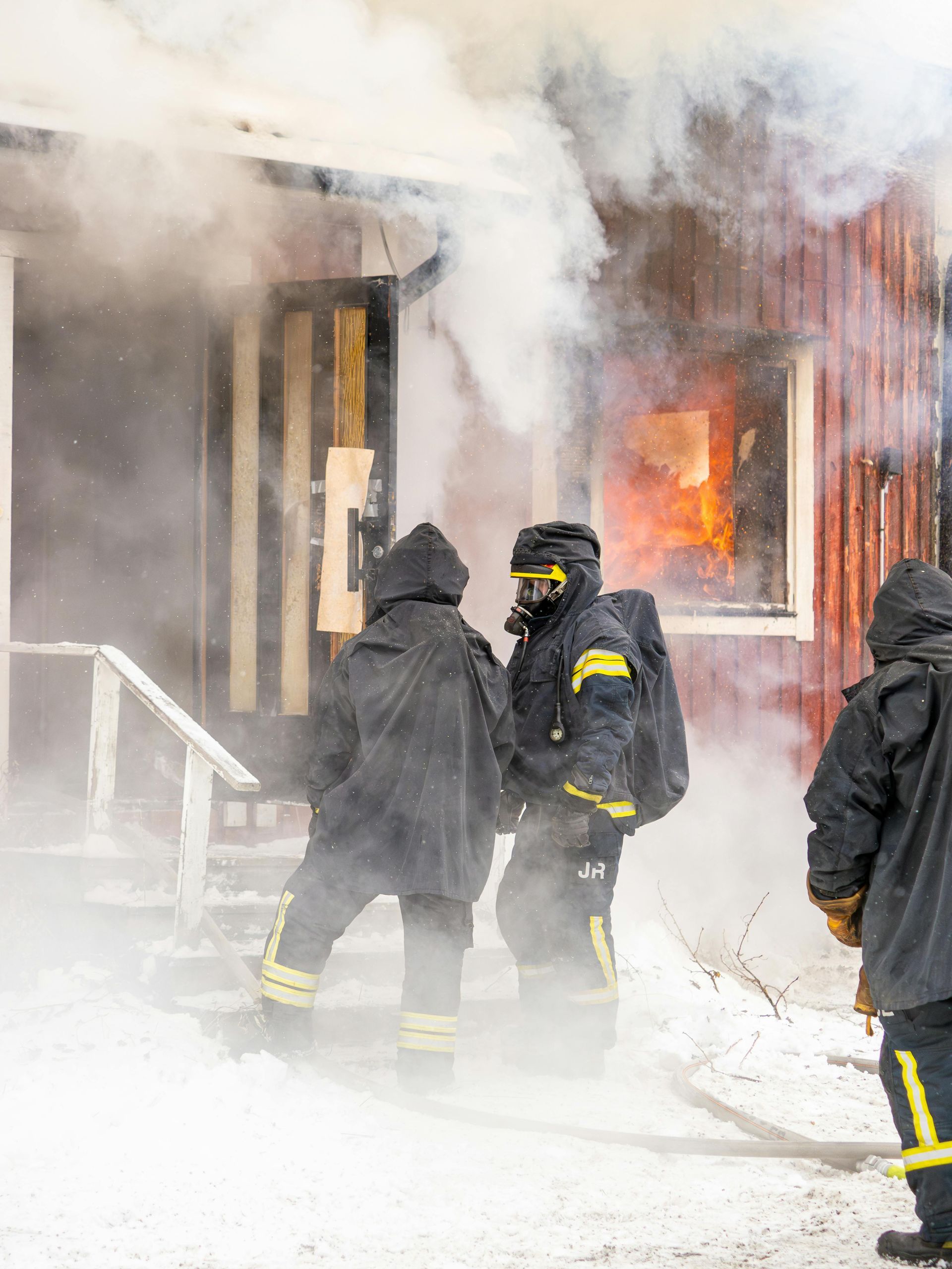 Three firefighters in full protective gear and masks stand in the snow outside a building emitting smoke and flames.