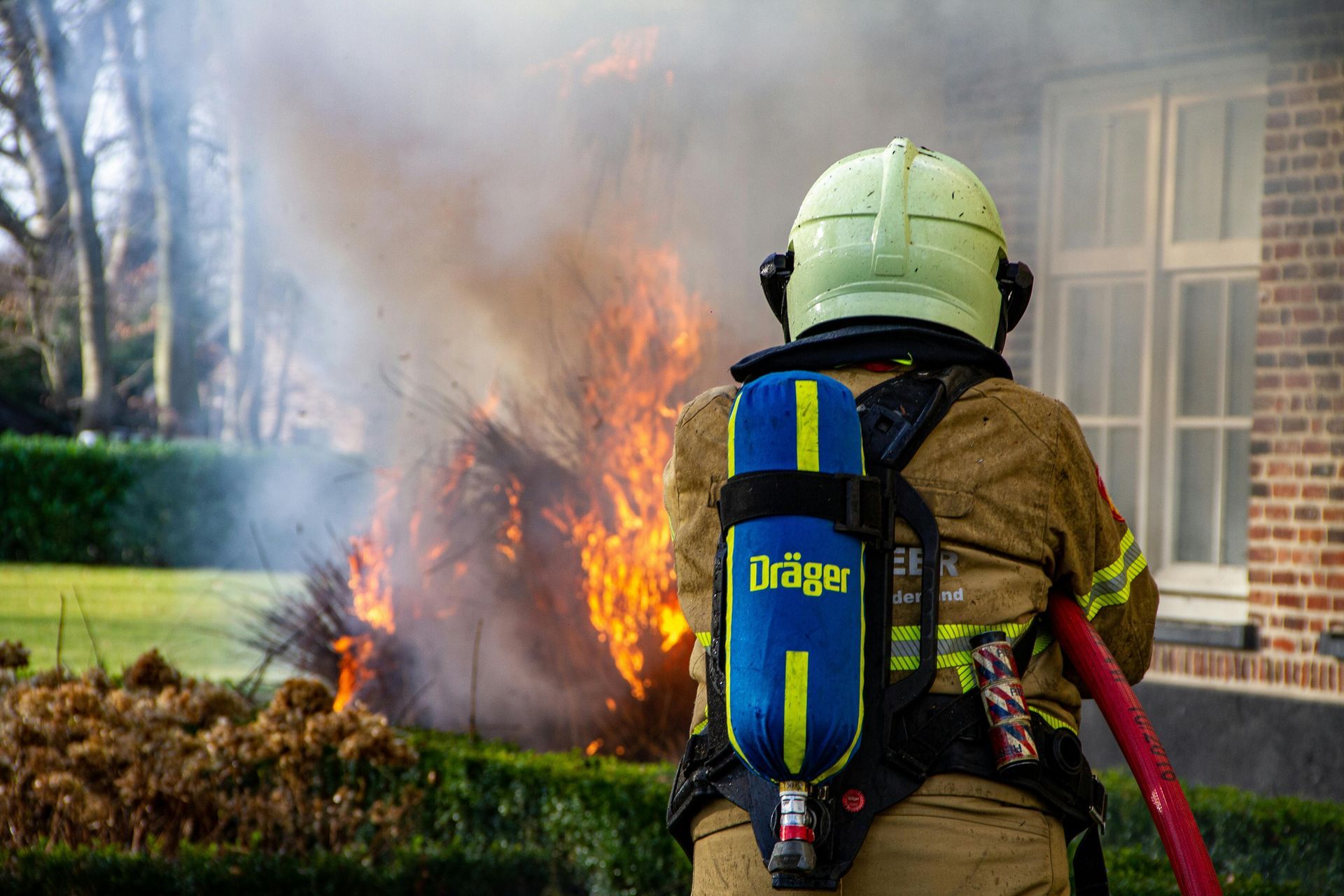 A firefighter in protective gear and a light-colored helmet sprays water on a burning hedge near a brick building.