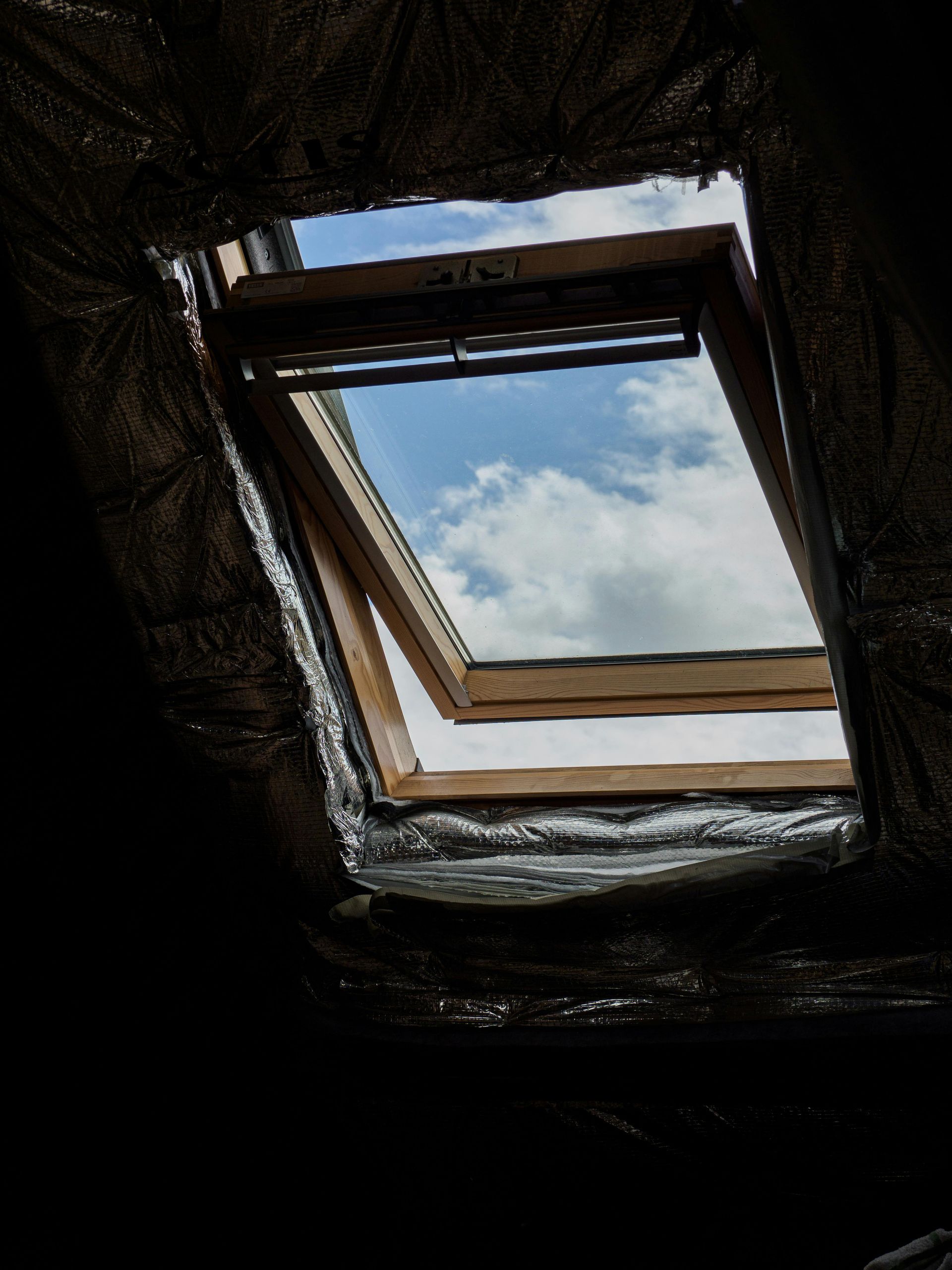 An open wooden skylight reveals a bright blue sky filled with white clouds, surrounded by reflective insulation.
