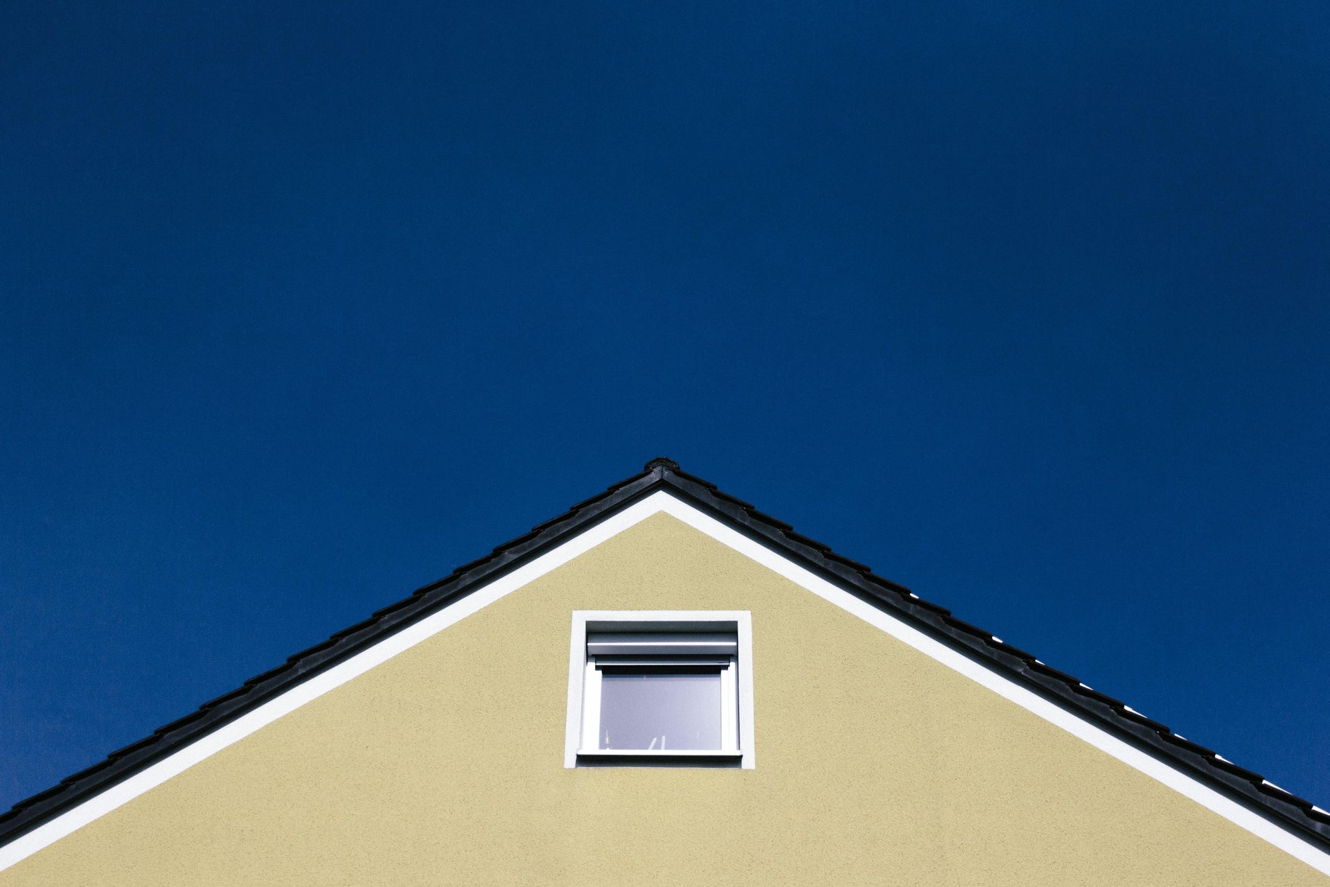 A beige triangular gable end of a house with a single window against a deep blue sky.