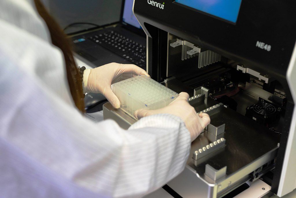 Persona in camice e guanti che posiziona un vassoio in uno strumento scientifico, vicino a un computer portatile.