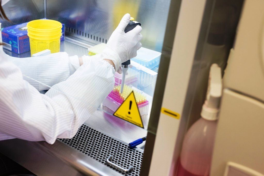 Scienziato in camice da laboratorio che utilizza una pipetta sotto una cappa di sicurezza, vicino a un cartello di pericolo e a un liquido.