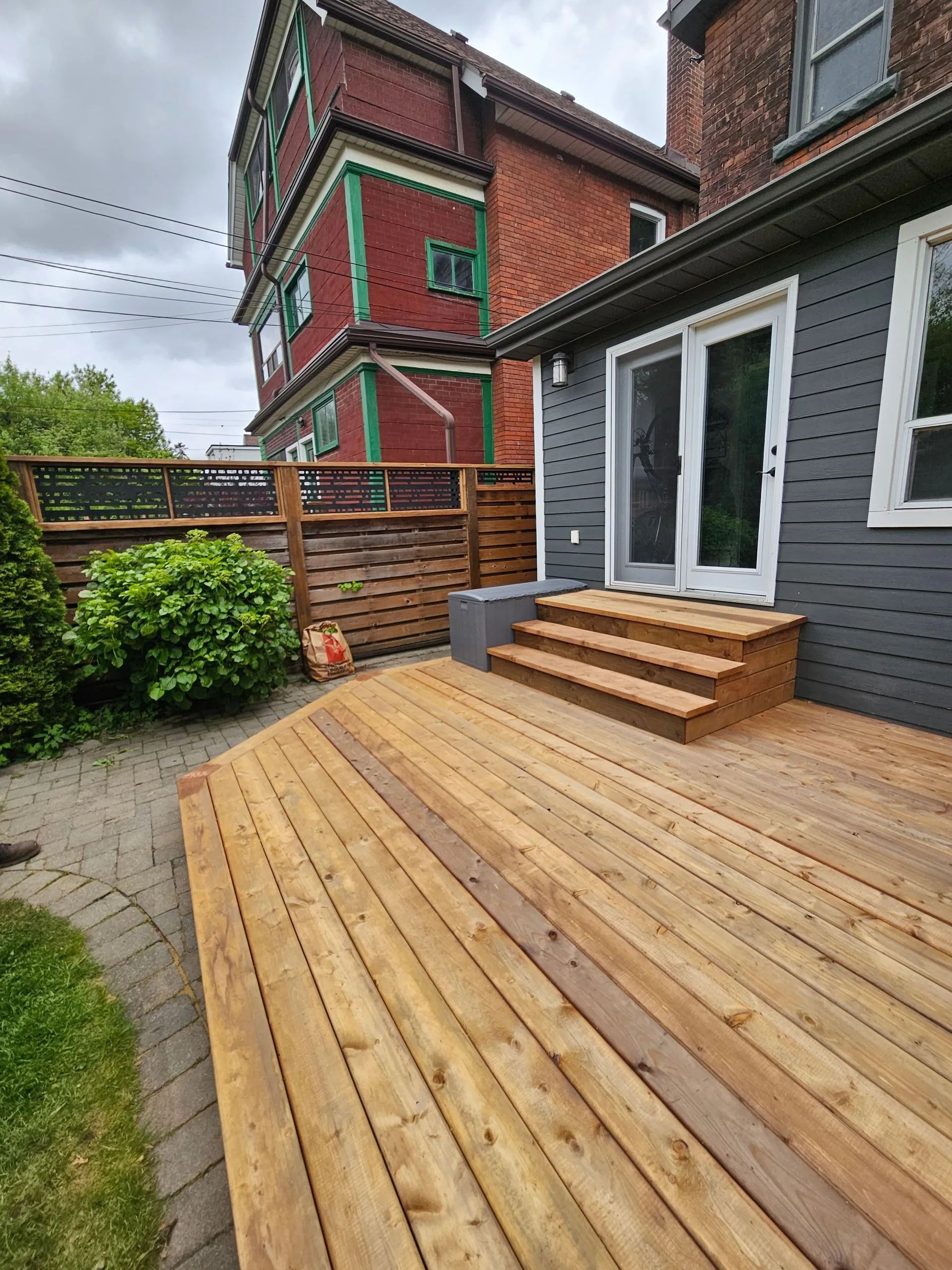 A new wooden deck with stairs leading to sliding glass doors on a dark gray house, next to a red building and fence.