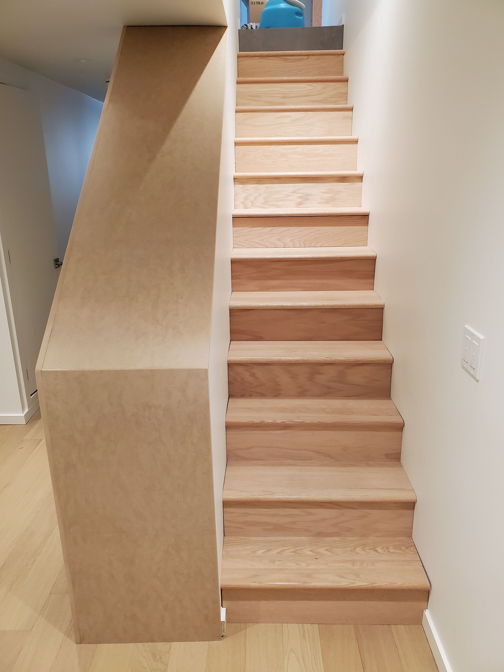 A straight flight of light-colored wooden stairs between a plain white wall and a tall, unfinished wooden pony wall.