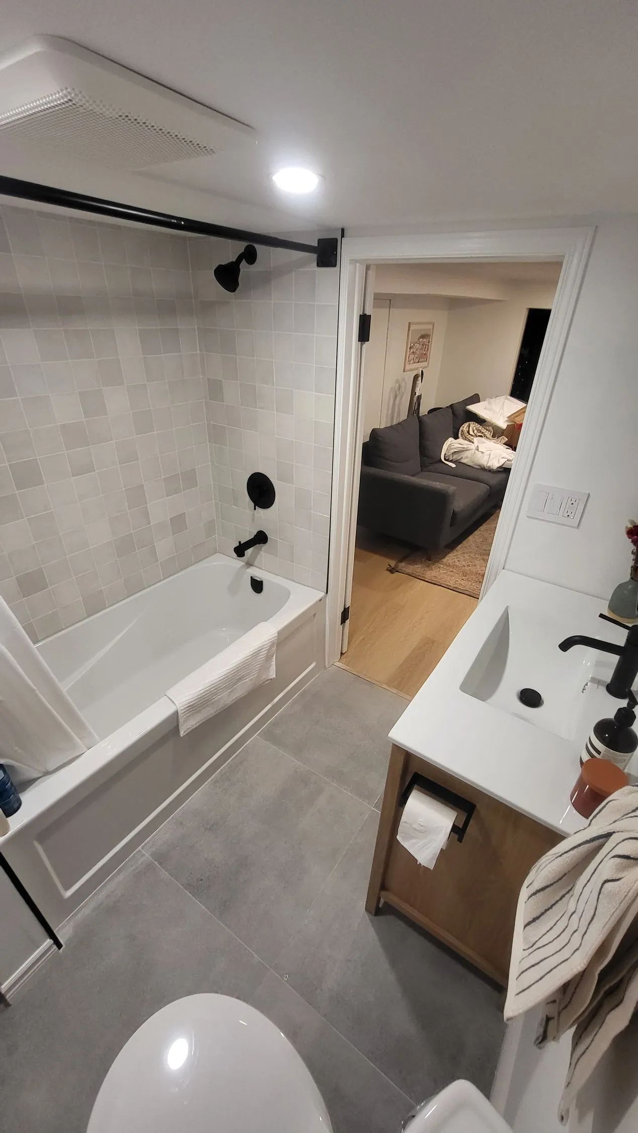 A modern bathroom featuring a white bathtub with black fixtures, gray floor tiles, and a wooden vanity, seen from above.
