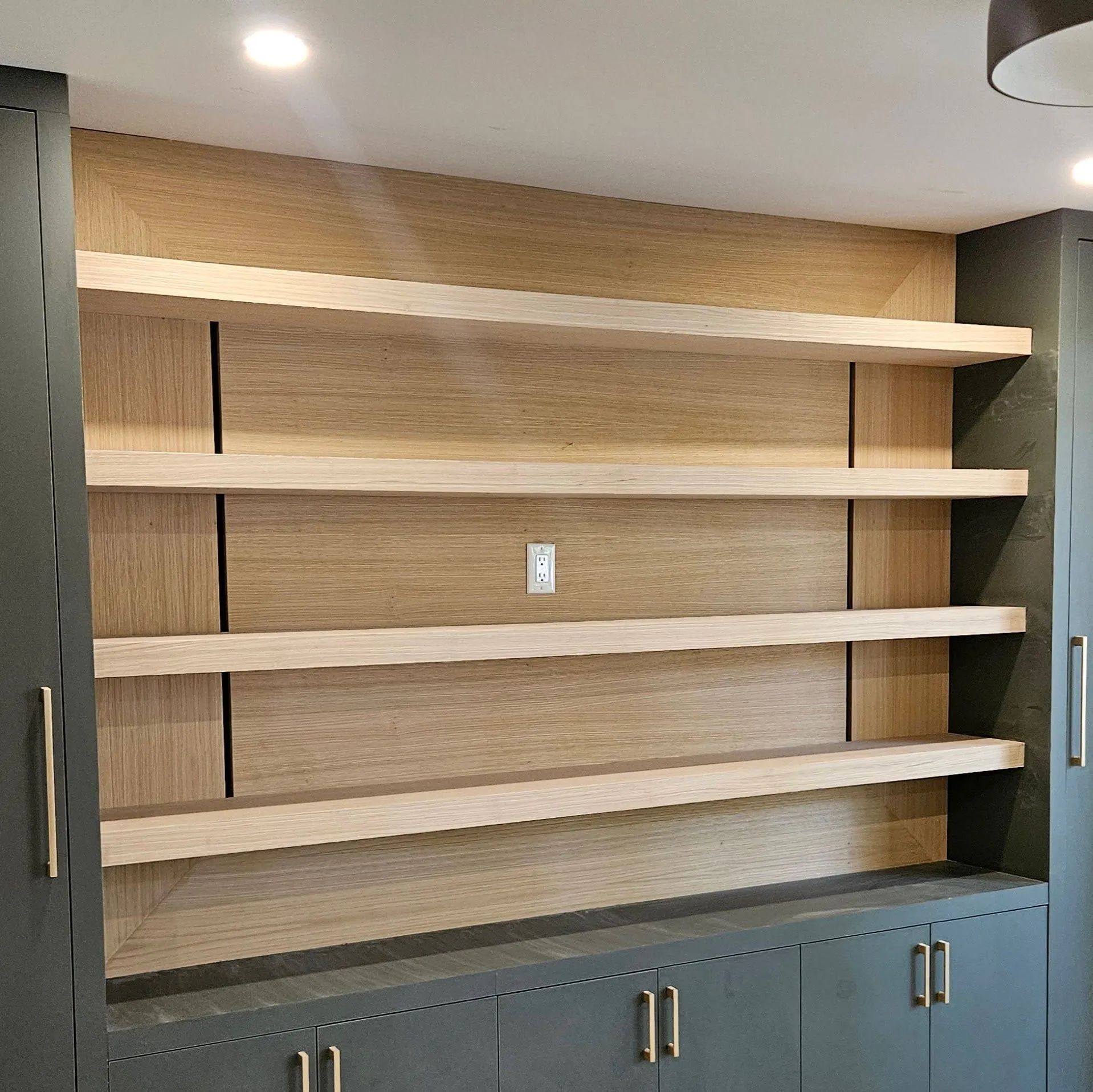 Modern wall unit featuring four light oak floating shelves centered against a wood-paneled wall above dark grey cabinets.
