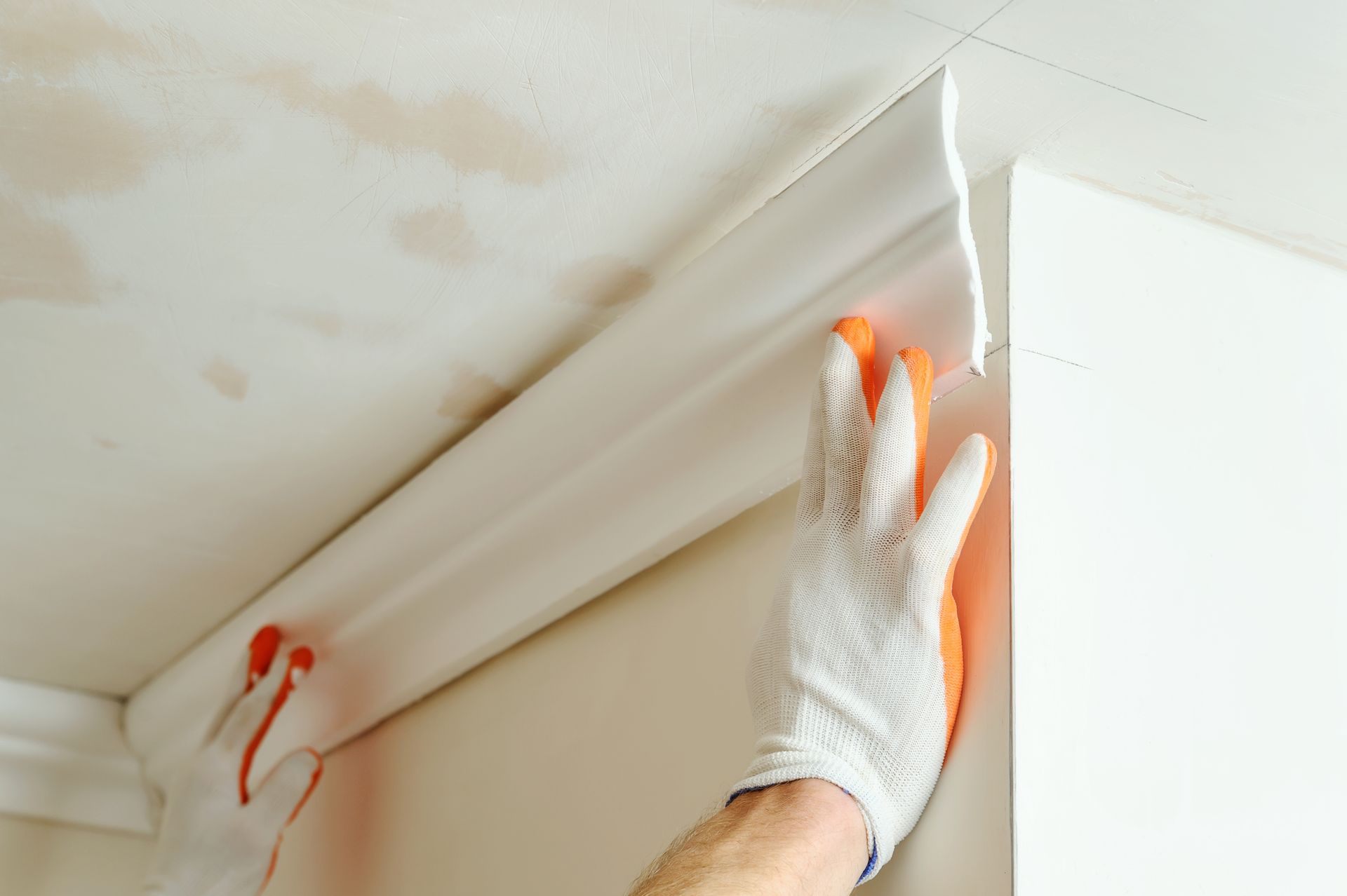 Worker installing crown molding on a ceiling corner, wearing gloves.