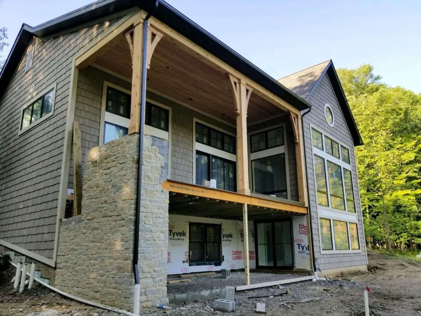 Two-story house under construction with stone columns, gray siding, and unfinished porch framing