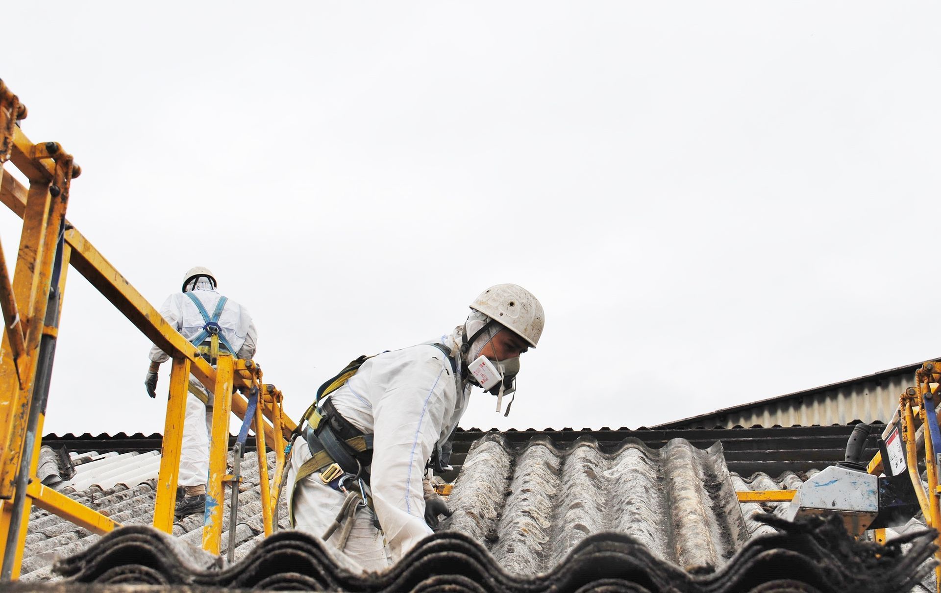 Worker in white safety gear crouches on a corrugated roof beside a yellow crane under an overcast sky