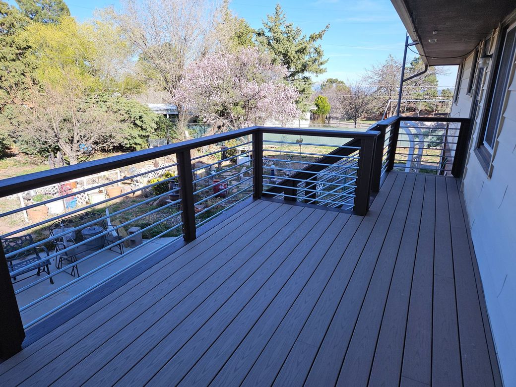 Wooden balcony deck with black railing overlooking trees and a hillside under a clear sky