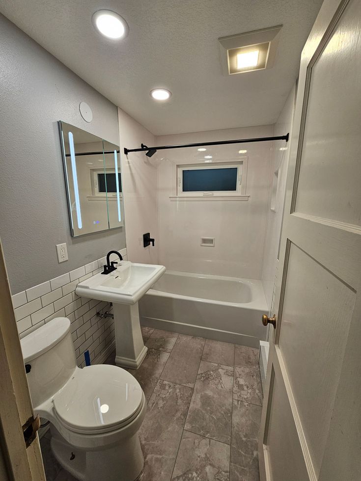 Compact white bathroom with toilet, pedestal sink, mirror, tub, and small window under bright ceiling lights