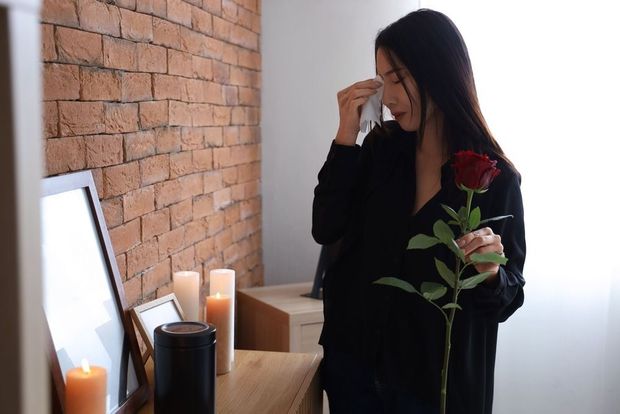 A person in black clothing holds a red rose while wiping away a tear near a shelf with candles and framed pictures.