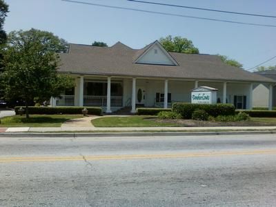 A one-story building with a wrap-around porch and white columns, fronted by a lawn and a sign reading