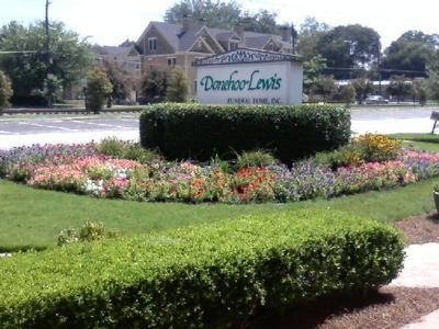 A stone sign for Donohoo-Lewis Funeral Home sits behind a lush flower bed and a manicured hedge on a sunny day.