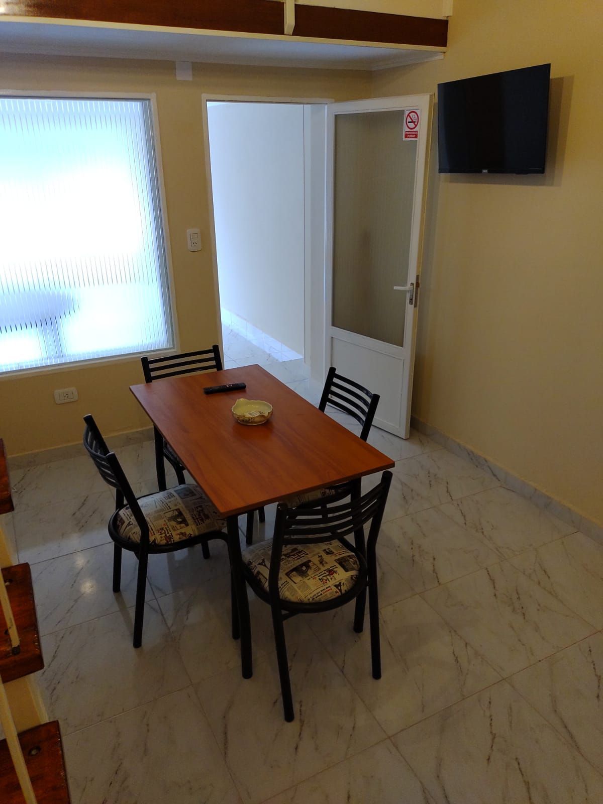 Comedor con mesa, cuatro sillas, TV en la pared, puerta a otra habitación, ventana.