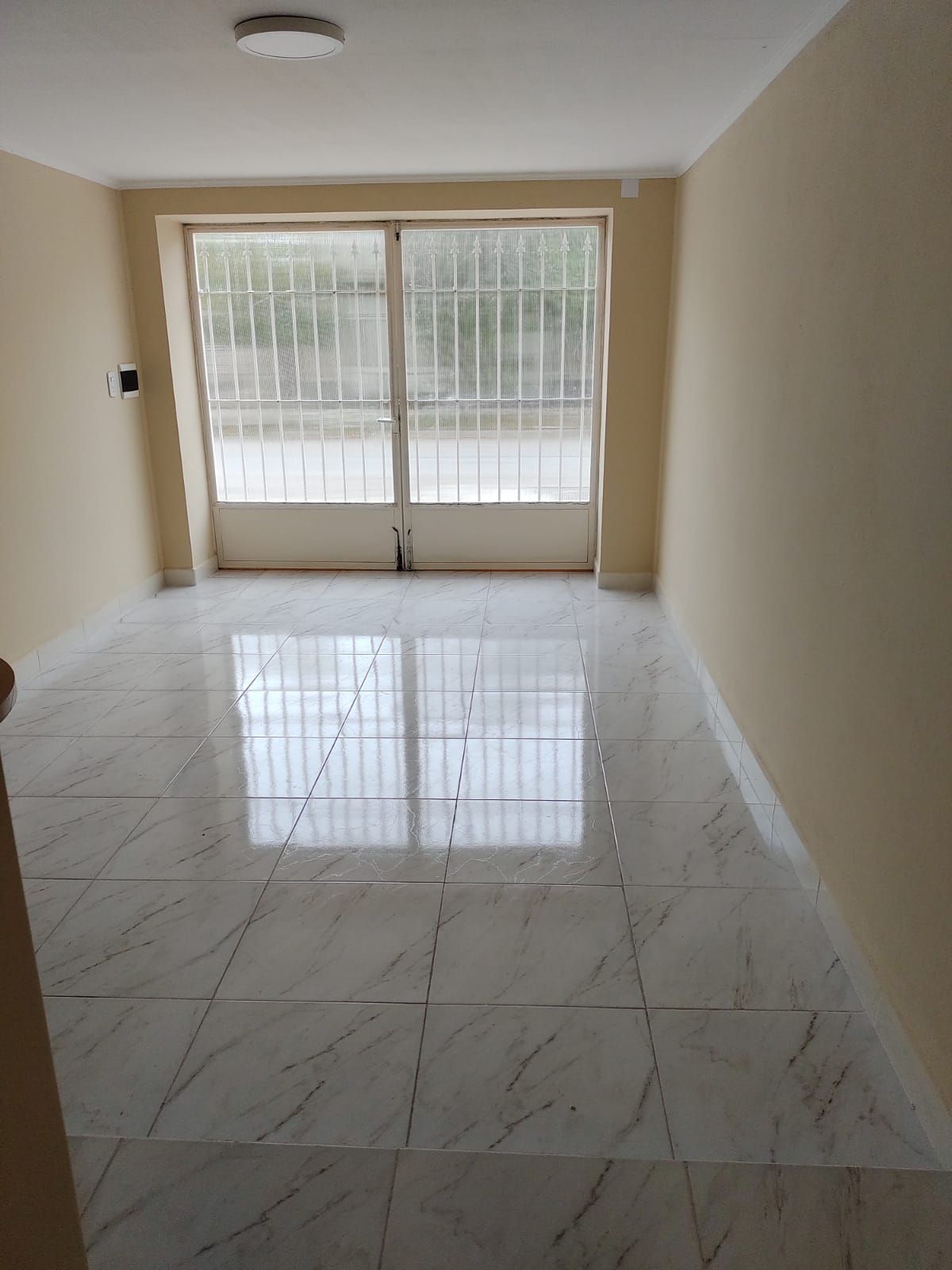 Habitación vacía con piso de baldosas claras, ventana grande con rejas de seguridad y paredes de color tostado.