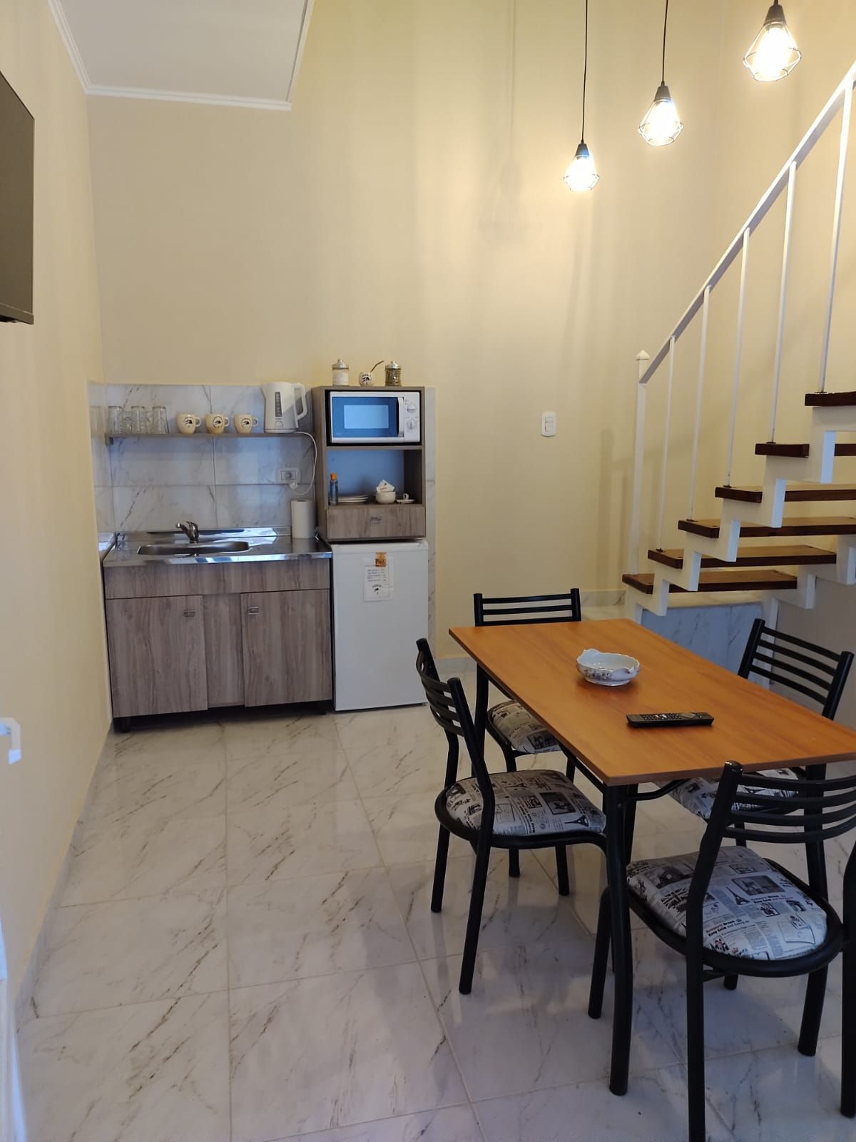 Cocina con comedor. Muebles de madera clara, refrigerador blanco, escalera. Mesa con sillas y luces de techo.