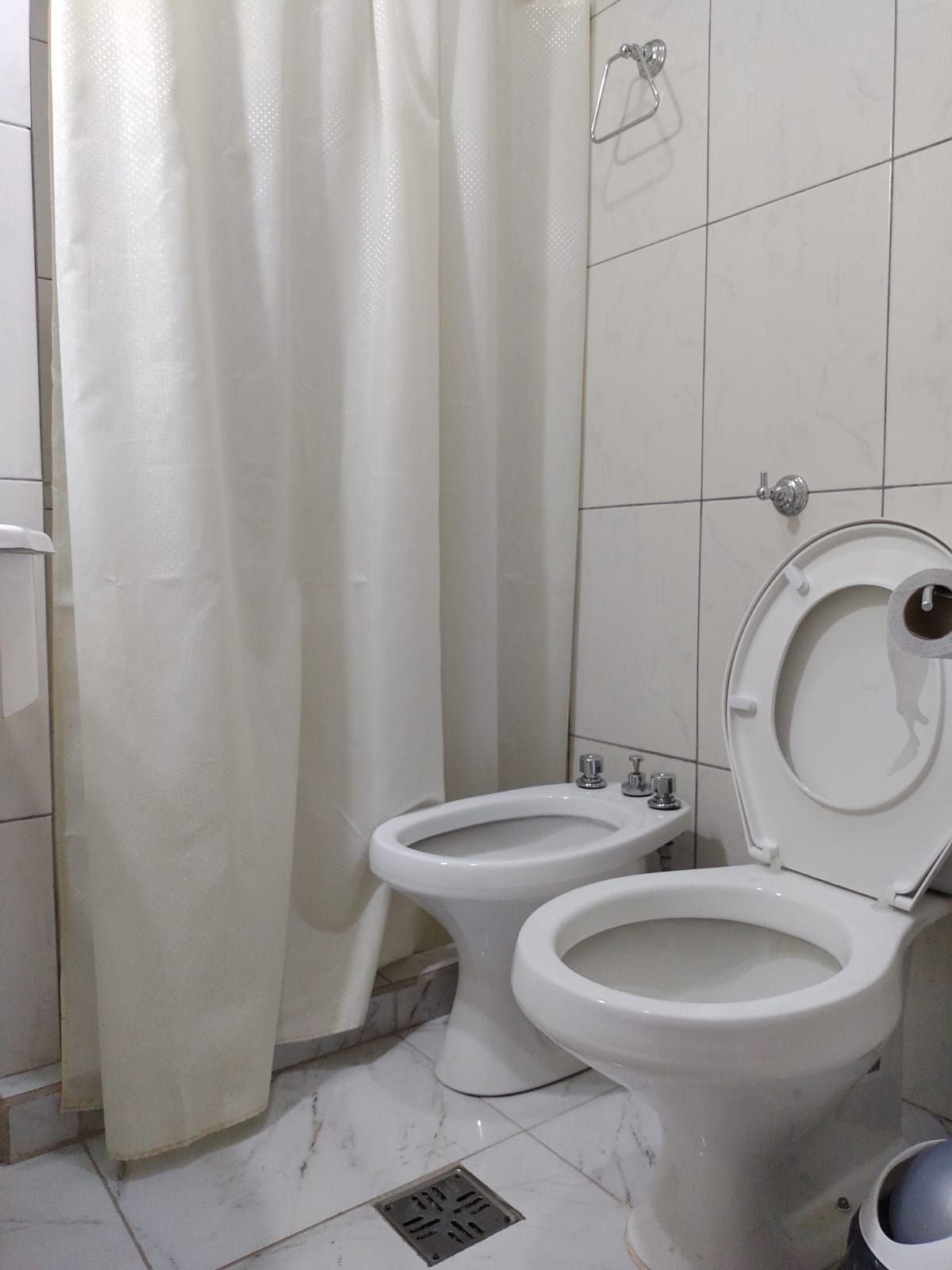 Baño blanco con inodoro, bidé y cortina de ducha contra paredes de azulejos blancos.