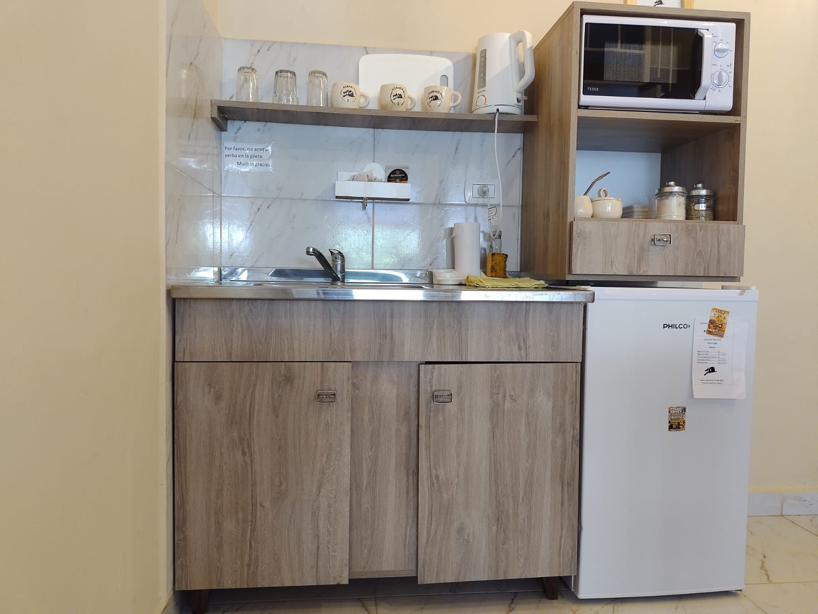 Pequeña cocina con microondas, refrigerador y fregadero; gabinetes de madera, paredes en tonos neutros.