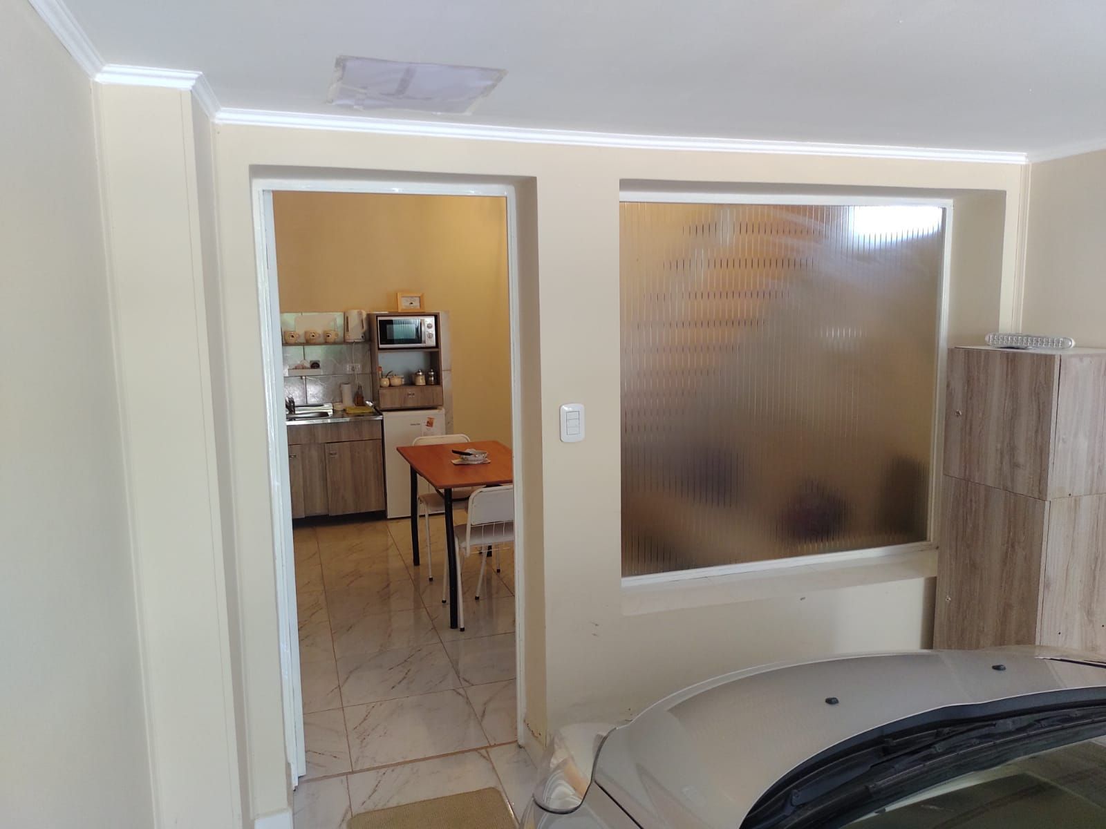 Interior con un coche aparcado, una cocina visible a través de una puerta y una ventana esmerilada. Paredes beige.