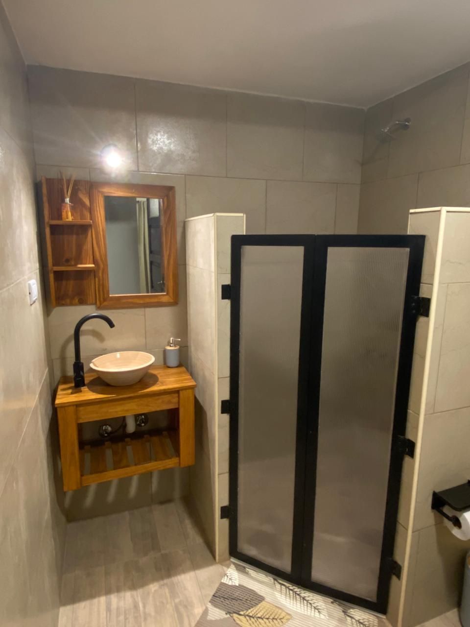Baño con tocador de madera, lavabo color tostado, grifo negro y puertas de ducha de vidrio.