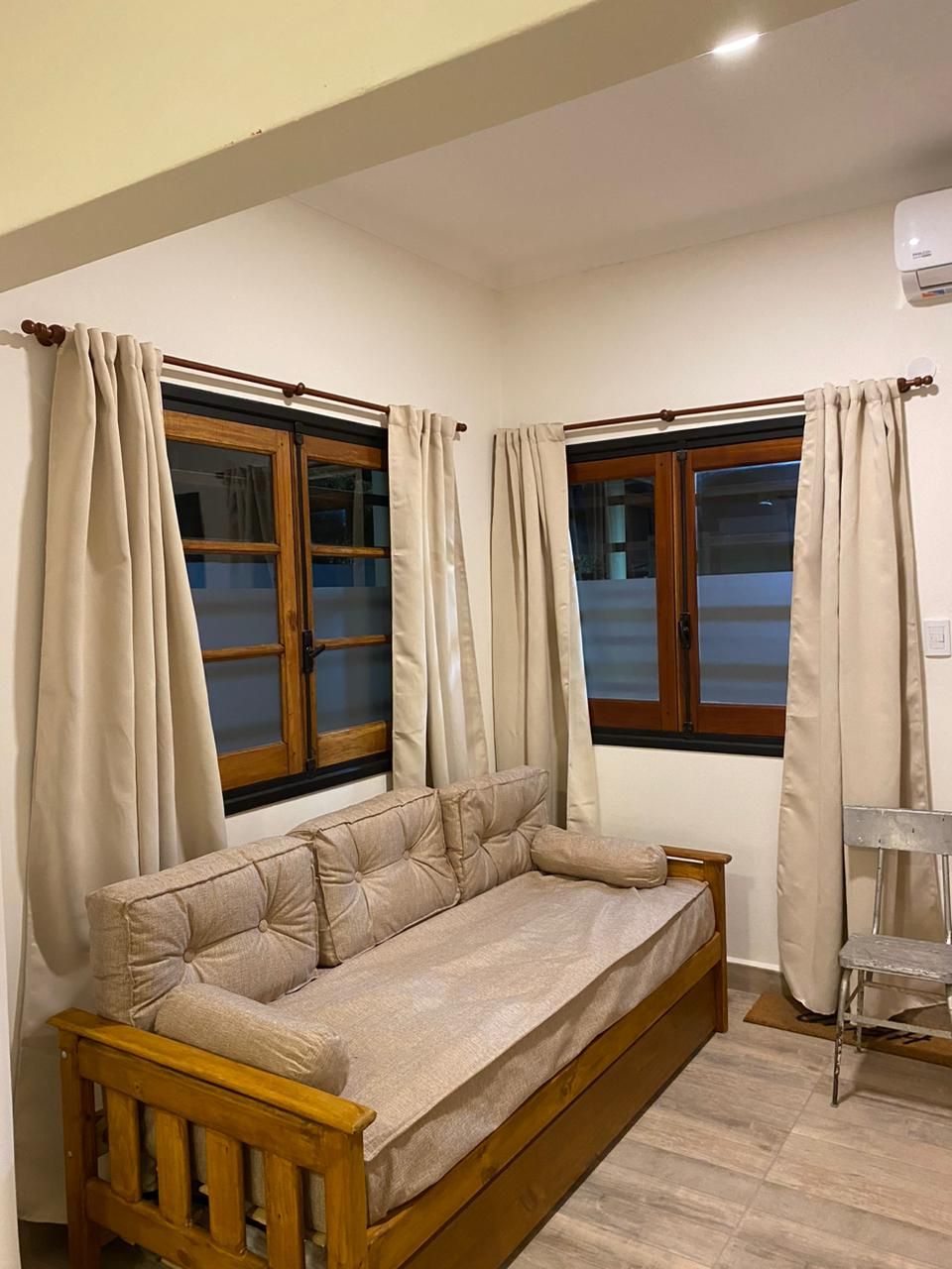 Acogedora sala de estar con un sofá cama de madera, dos ventanas con cortinas y una pequeña silla de metal.