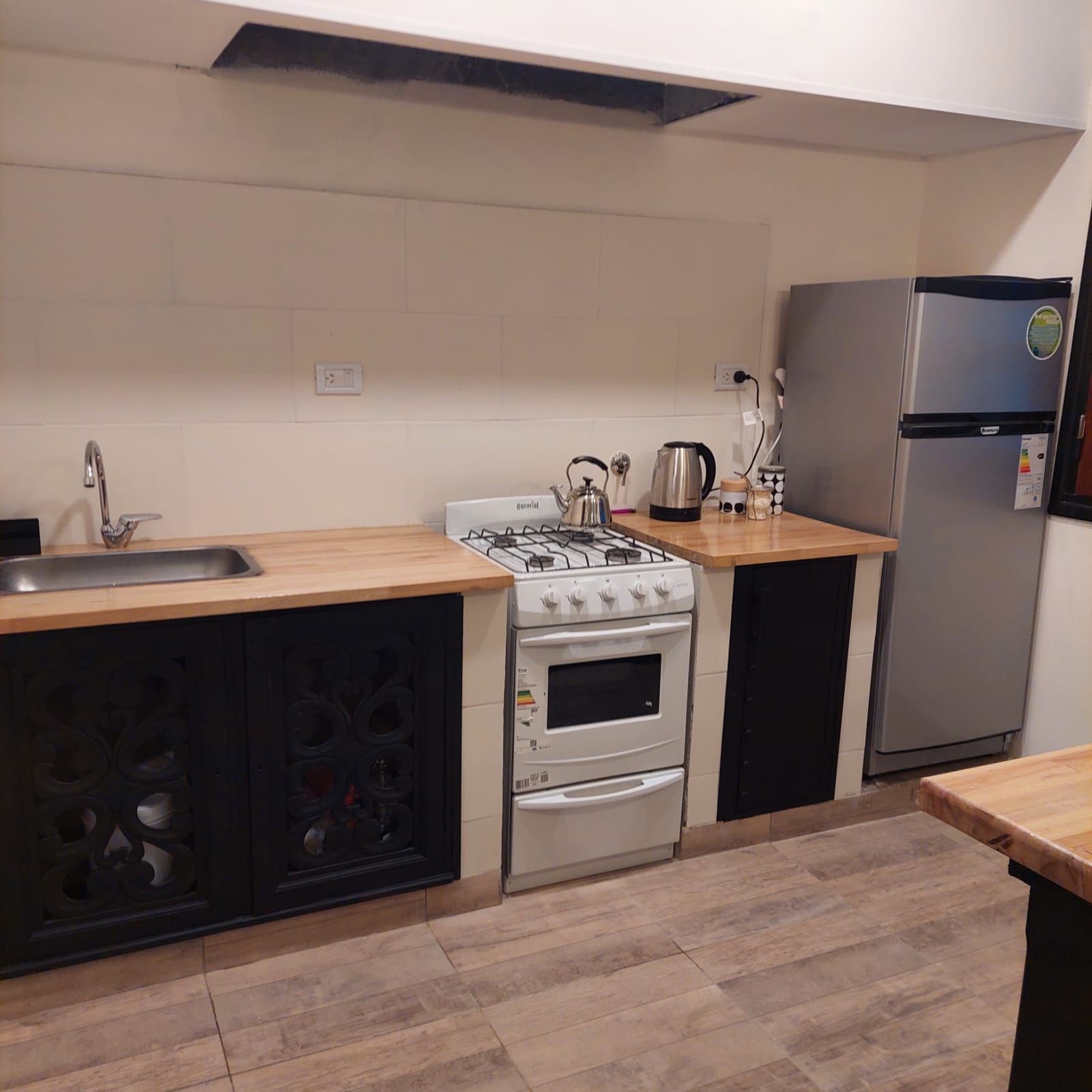 Pequeña cocina con gabinetes negros, encimeras de madera y un refrigerador plateado.